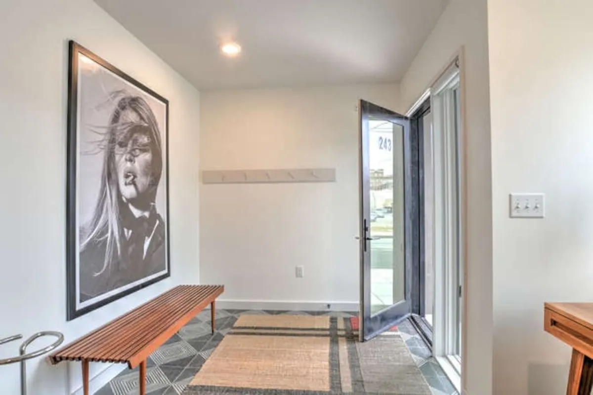 A stylish entryway featuring a modern wooden bench set on a geometric patterned rug, with a large black and white portrait as a focal point and an open door suggesting a transition into the home. The space is bright, inviting, and designed for welcoming guests or pausing when coming and going.