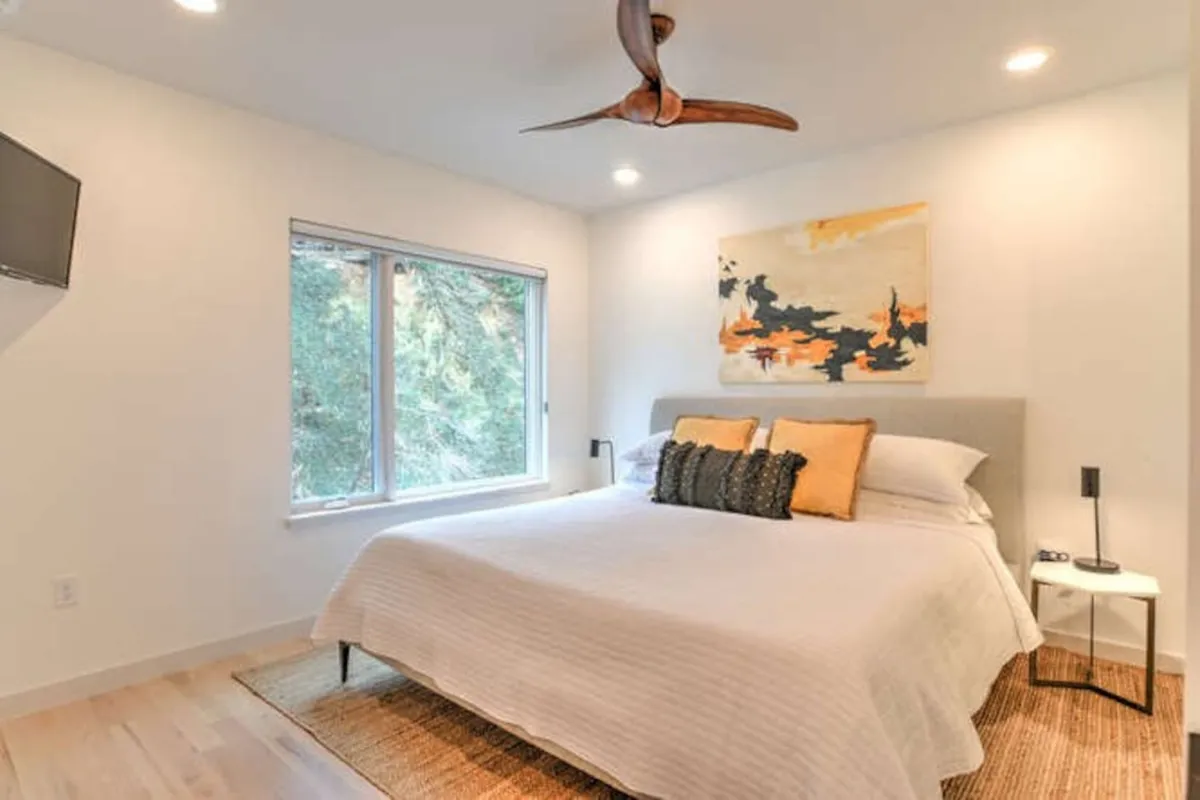 A serene king bedroom featuring a large bed with plush bedding, a window letting in natural light, tasteful artwork above the headboard, and a ceiling fan overhead. The overall ambiance is calm and inviting, ideal for rest and relaxation.