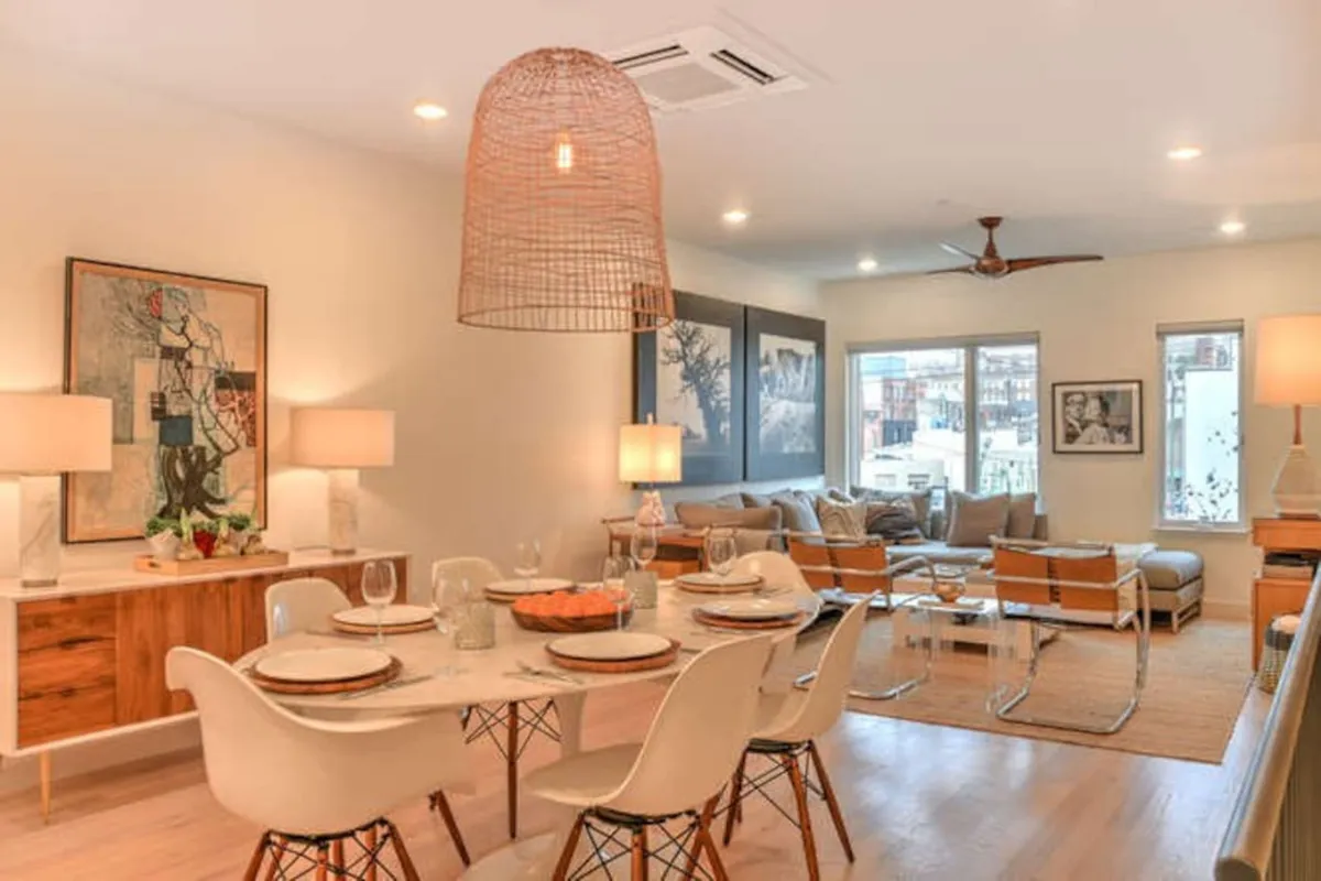 This image presents a dining table set for a meal, surrounded by comfortable seating and accented by a large pendant light overhead. Visible artwork and stylish decor establish a warm, inviting mood perfect for gatherings. The background reveals a seamless flow into a cozy seating area, exemplifying open-concept design.