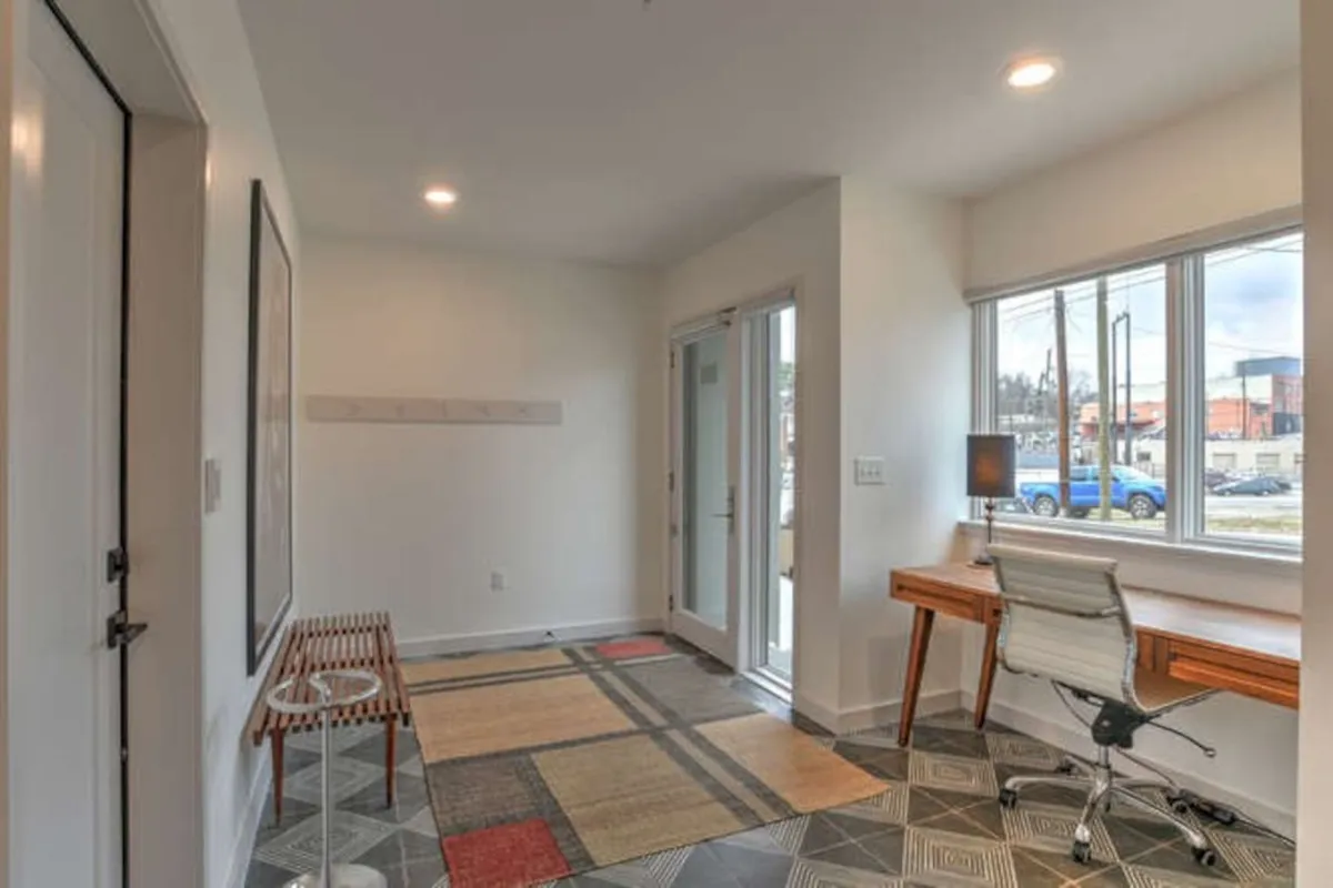 An inviting workspace featuring a desk with a comfortable chair positioned beside large windows. The room is accented by a colorful rug and vibrant artwork, with sunlight filtering through, creating a lively yet serene atmosphere. The view outside the windows subtly suggests a connection to the broader city environment.