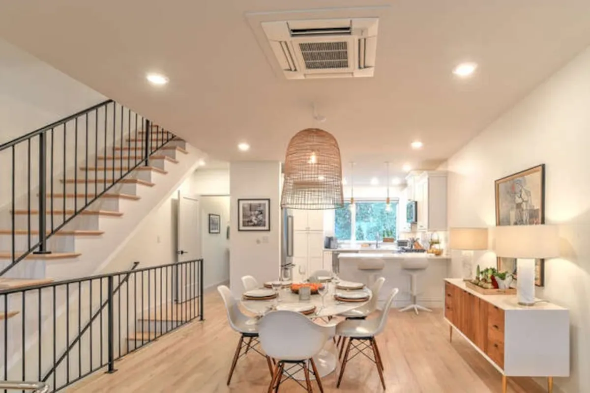 This image showcases a contemporary dining area with a decorative centerpiece on the table, a hanging light fixture, a staircase leading to the upper floor, and a kitchen area visible in the background. The scene feels open, modern, and emphasizes the vertical flow of the home.