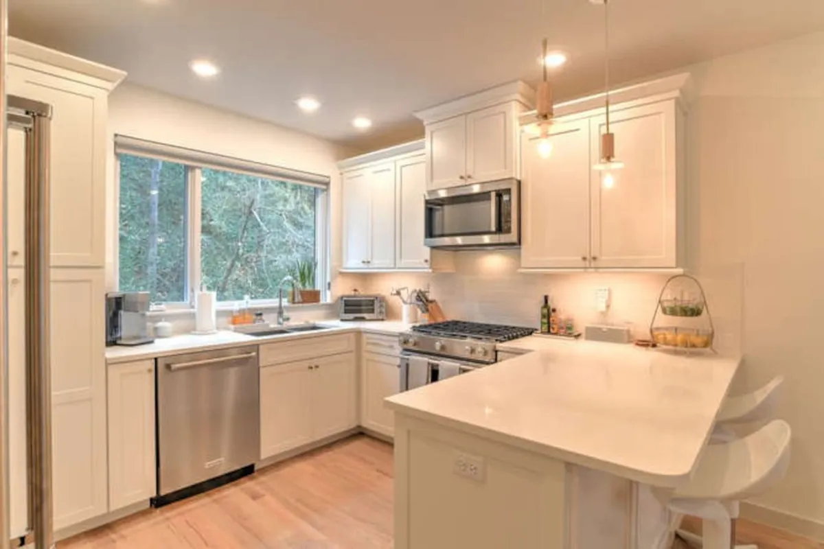 A bright, modern kitchen with a large window that offers a glimpse of lush greenery outside. The scene features a sleek kitchen island, modern cooking appliances, and minimalist cabinetry, creating an inviting environment perfect for cooking and socializing. Natural light streams in, forging a strong visual connection to the outdoors and hinting at the property's residential setting within a vibrant neighborhood.