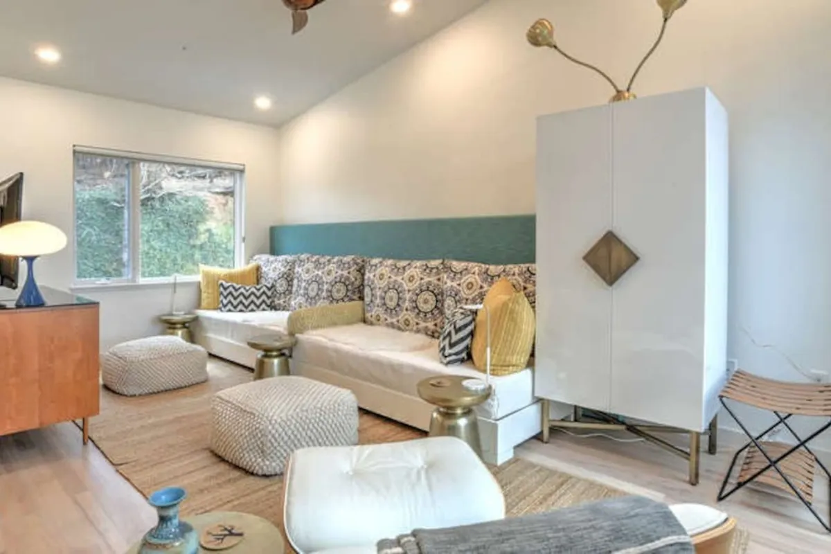 A stylish living area featuring a cozy sofa with patterned cushions, gold accent tables, large windows with a view of greenery, and a modern light fixture. The room is filled with natural light and offers a glimpse of the outdoors, connecting interior comfort with the world beyond.