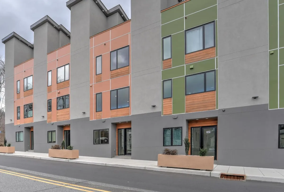This exterior image showcases colorful façades, large windows, and inviting wooden accents along a contemporary street. Concrete planters and the open sidewalk set the scene for an active, social neighborhood ambiance, suggesting a vibrant community atmosphere just outside the door.