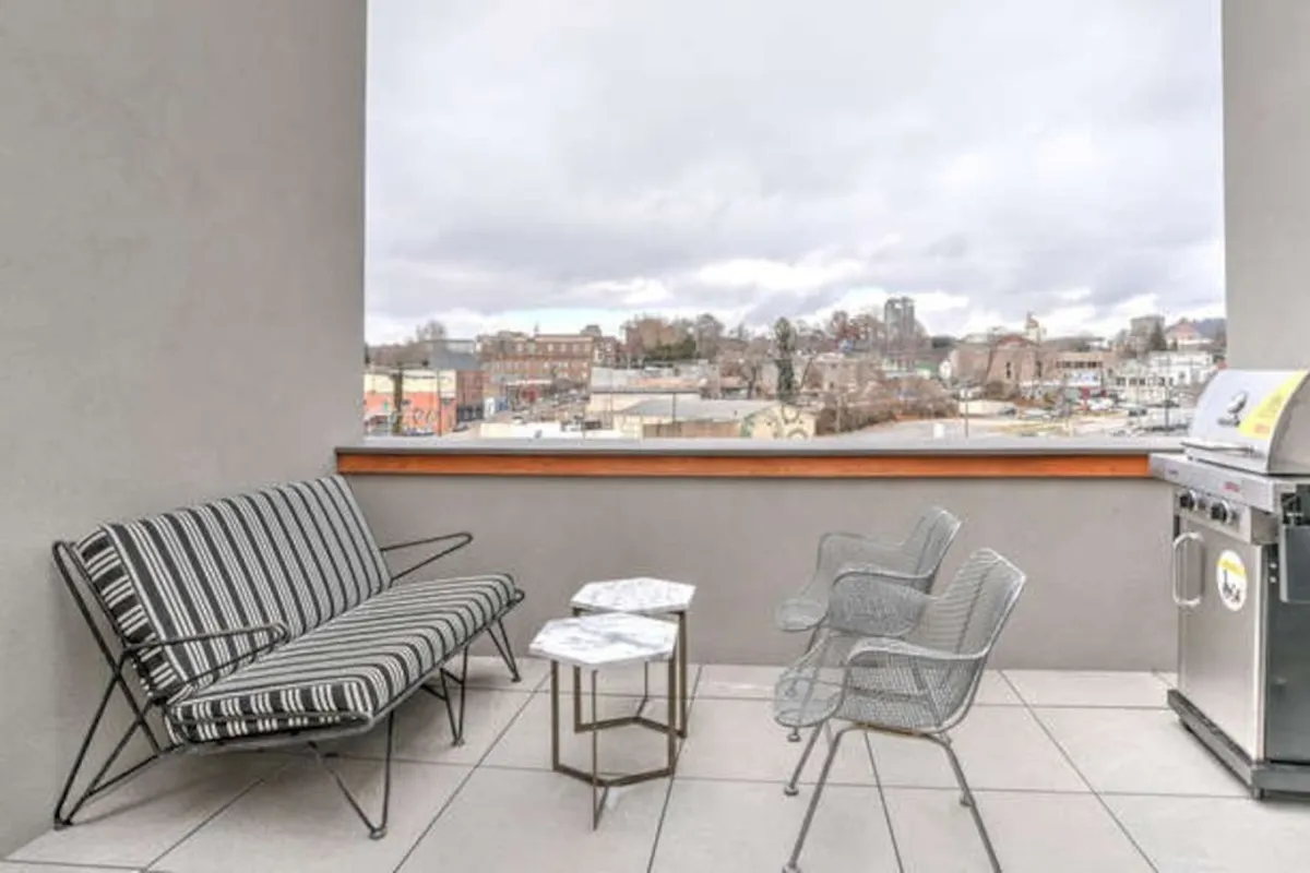 This exterior image showcases a stylish rooftop patio furnished with a striped sofa and a marble table, positioned to take in panoramic city views. A grill is also present, suggesting opportunities for outdoor cooking and gatherings. The urban skyline forms an inviting backdrop, creating an atmosphere that is both casual and elevated.